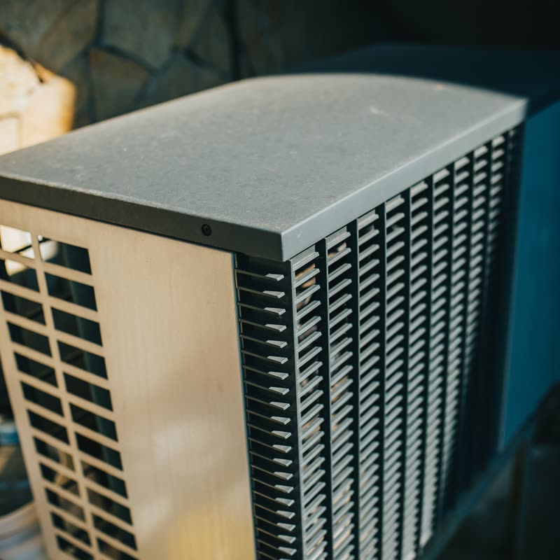 Lavt sollys på siden og lamellerne af en udedel til aircondition, der står op ad en stenmur i en have